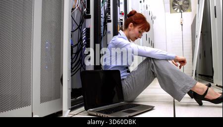 Sitzende Frau in blauem Hemd, die mit Kabeln zwischen Serverschränken im Rechenzentrum einen Laptop überprüft Stockfoto