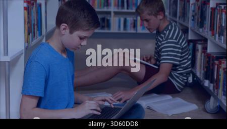 Schreibjunge in blauem T-Shirt mit silbernem Laptop im Gang der Schulbibliothek, mit Tablet, offenem Lehrbuch Stockfoto
