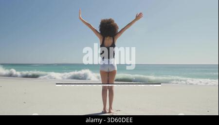 Stehende Frau, die Arme am Strand hochhebt, in schwarzem Tanktop und Shorts, Wellen, Artefaktüberquerung Stockfoto