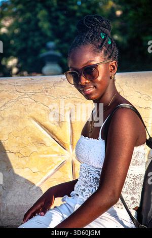 Junge afro-kubanische Frau posiert in weißer Kleidung und Sonnenbrille auf den Straßen von Santiago de Cuba, Kuba. Model sitzt auf einem Stuhl bei Sonnenuntergang. Stockfoto