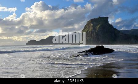 Piha Beach, Neuseeland, hat Wellen, die am Ufer mit einem dunklen Felsen im Vordergrund und einer großen, grünen Landzunge unter einem teilweise bewölkten Himmel zusammenbrechen. Stockfoto