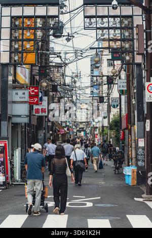 Die Menschen laufen in einem lebhaften Viertel von Osaka eine geschäftige, enge Straße entlang, die von verwickelten Versorgungsleitungen, alten Schildern und einzigartiger Architektur gesäumt ist. Stockfoto