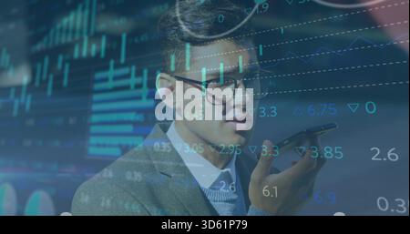 Sprechender asiatischer Geschäftsmann mit Smartphone am Schreibtisch, der überlagerte Diagramme in Anzug und Brille ansieht Stockfoto