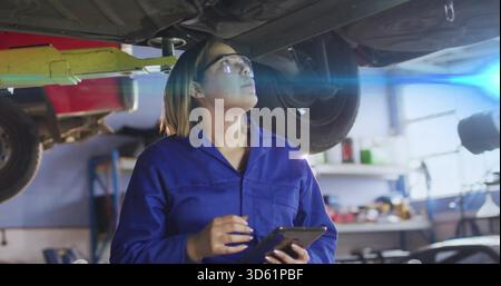 Inspektion einer Mechanikerin in blauen Overalls mit Schutzbrille, die das Auto im Aufzug mit einem Tablet überprüft Stockfoto