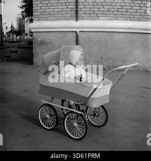 UdSSR, Ukrainische SSR, Slawjansk, 1970er Jahre Ein bezauberndes, abendfüllendes Porträt eines Jungen, der in einem klassischen Kinderwagen aus der Sowjetzeit sitzt. Das Kind trägt eine Motorhaube und leichte Kleidung, hält ein Spielzeug in den Händen und sieht neugierig zur Seite. Der Kinderwagen verfügt über große Speichenräder und ein Retro-Design. Es ist auf einem gepflasterten Bürgersteig neben einer Backsteinmauer geparkt. Dieses Schwarzweiß-Archivfoto fängt die Ästhetik des Kindheits- und Alltags in der ruhigen Donbass-Region ein. Stockfoto