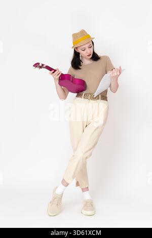 Die junge weibliche Ukulele-Künstlerin hält Ukulele und Noten und studiert intensiv musikalische Noten und Akkorde. Blick auf die ganze Länge der seriösen Frau, stilvoll getragen Stockfoto