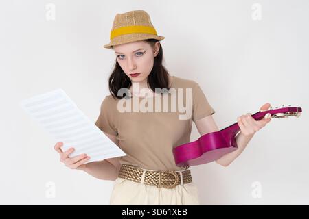 Porträt einer jungen Ukulele-Spielerin hält Ukulele und Noten in der Hand und blickt in die Kamera. Junge und schöne Frau der kaukasischen eth Stockfoto