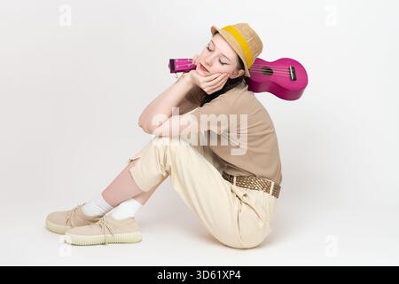 Sorglos aussehender Musiker sitzt mit geschlossenen Augen auf dem Boden, Ukulele über die Schulter geschlungen, das Kinn in der Hand. Eine junge Instrumentalistin im lässigen Clo Stockfoto