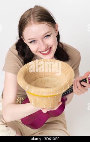 Fröhlicher, lächelnder Musiker mit Ukulele, der Strohhut hält, um Spenden und Trinkgelder zu erhalten, Geld und andere Wertzeichen zu sammeln. Sie Stockfoto