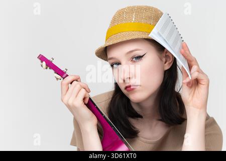 Nahaufnahme Portrait müder und emotional erschöpfter Ukulele-Künstler mit Ukulele und Noten in den Händen. Frau, die in die Kamera schaut und nachdenkliche Ausdrücke vermittelt Stockfoto
