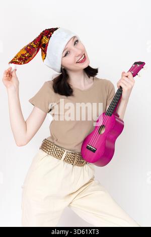 Die junge Musikerin mit rotem Weihnachtsmütze und Ukulele in der Hand posiert spielerisch und lenkt den Blick auf die Kamera. Die schöne Frau flirtet mit dem Flirtatio Stockfoto