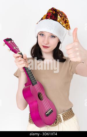 Eine junge Musikerin mit rotem Weihnachtsmann-Hut, Ukulele in der Hand und Daumen hoch. 18 Jahre alte kaukasische Ukulele-Künstlerin, die in die Kamera schaut Stockfoto