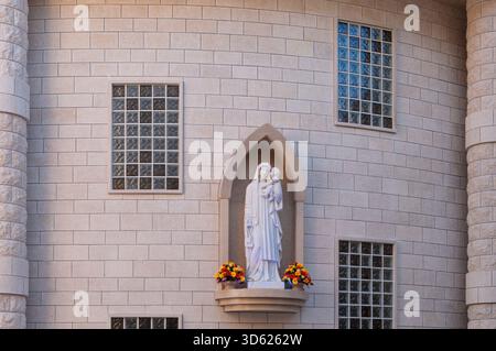 Eine Statue von Maria, die ein Baby hält, steht auf einem Felsvorsprung eines Gebäudes. Die Statue ist von Blumen umgeben und steht vor einem Fenster Stockfoto