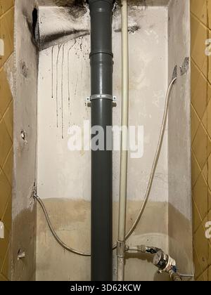 Der Schrank zeigt einen schweren Zerfall durch chronischen Wasserverlust. Schwarze Flecken und feuchter Putz umgeben ein vertikales PVC-Ablaufrohr. Ausfall der Sanitärinfrastruktur und dringender Wartungsbedarf. Stockfoto