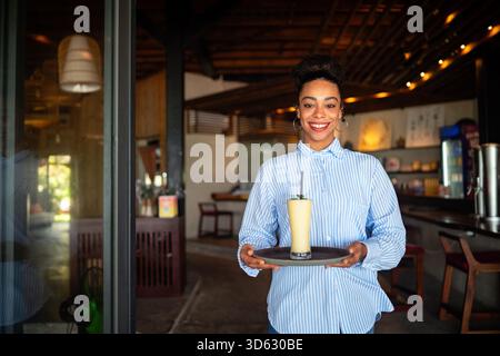 Junge lächelnde afroamerikanische Kellnerin, die Fruchtsaft und Smoothie serviert, während sie in einem Café arbeitet. Stockfoto