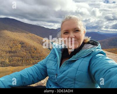 Eine lächelnde Frau sitzt auf einem Felsen auf der Spitze eines Berges und bewundert die atemberaubende Herbstlandschaft voller leuchtender orange und gelber Bäume. Der Himmel Stockfoto