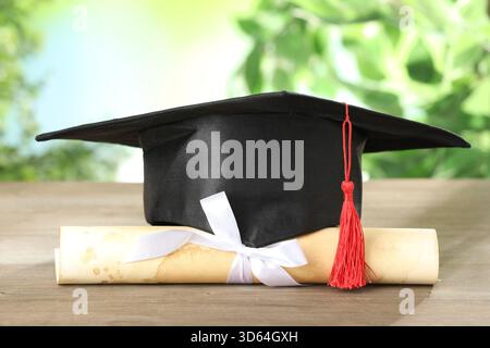 Graduierungskappe und Diplom auf Holztisch vor verschwommenem Hintergrund, Nahaufnahme Stockfoto