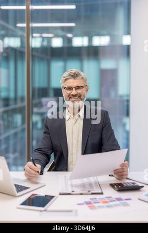 Reifer Geschäftsmann sitzt am Schreibtisch, hält Dokumente und Stift in der Hand, lächelt in die Kamera mit einem modernen Hintergrund im Büro und repräsentiert den beruflichen Erfolg und das Unternehmensumfeld Stockfoto