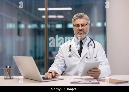 Professioneller, männlicher Arzt mit weißem Mantel und Stethoskop, der an einem modernen Schreibtisch sitzt, mit einem Laptop arbeitet und ein digitales Tablet hält, um medizinisches und medizinisches Fachwissen bereitzustellen Stockfoto