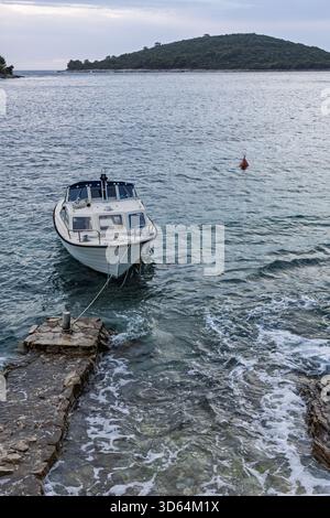 Ein kleines weißes Boot ist an einen Steinsteg im ruhigen blauen Wasser gebunden, mit einer bewaldeten Insel in der Ferne. Eine ruhige Küstenszene, die auf Reisen hindeutet, le Stockfoto