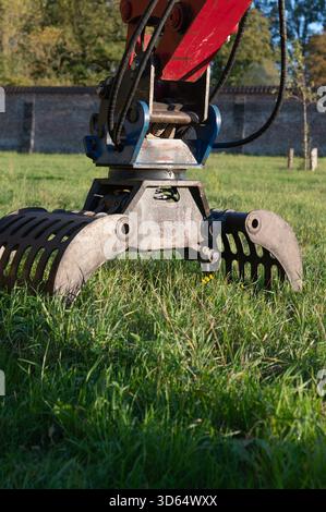 Roter Baggergreifer über grasbewachsenem Feld, vertikaler Winkel mit Ausleger und Hydraulikzylinder, Maschinen über Rasen in der Nähe von Ziegelwänden, Herbstlicht Stockfoto