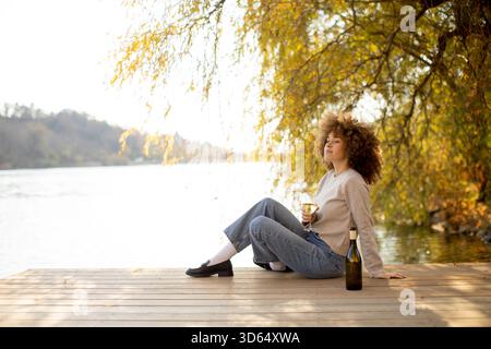 Eine junge Frau entspannt sich auf einem hölzernen Dock neben einem ruhigen Fluss und genießt einen Drink, während sie von leuchtenden Herbstfarben umgeben ist. Stockfoto
