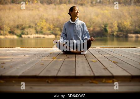 Eine Koreanerin sitzt friedlich auf einer Holzterrasse, meditiert neben einem ruhigen See, der von Herbstlaub umgeben ist, und genießt den Moment. Stockfoto