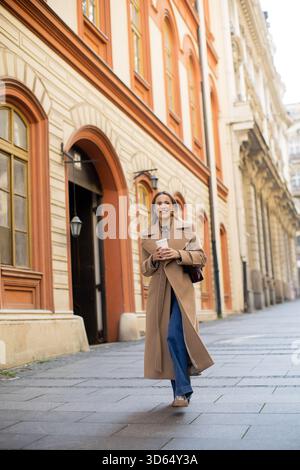 Eine junge Frau schlendert durch eine malerische Straße in einer Stadt, trägt einen stilvollen beigefarbenen Mantel und hält eine Kaffeetasse in der Hand. Helle Gebäude umgeben sie, Crea Stockfoto