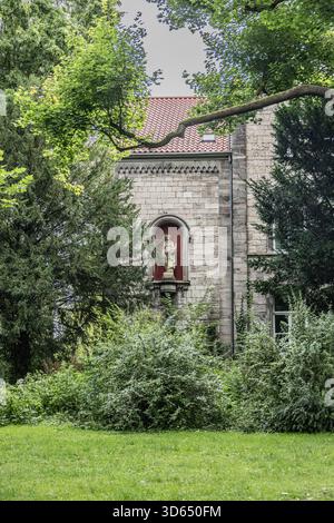 Steinmauer mit religiöser Statue in einer Nische, umgeben von dichtem Grün und Bäumen in Deutschland Stockfoto
