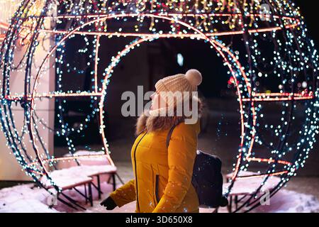 Die junge Touristin in Strickmütze schaut sich festliche Weihnachtsdekorationen an. Weihnachtsmarkt, Nachtbeleuchtung. Stockfoto