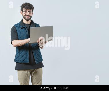Ein lächelnder Bärtiger, der Laptop benutzt und die Kamera isoliert betrachtet. Hübscher junger Mann mit Brille, Laptop und Blick in die Kamera Stockfoto