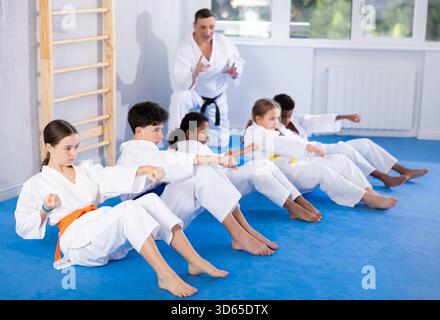 Gruppe multinationaler Kinder im Kimono trainieren Bauchmuskeln und machen gleichzeitig Schläge während Karate- oder Judo-Trainings Stockfoto