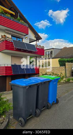 Wohngebäude mit Solarpaneelen auf den Balkonen. Im Vordergrund befinden sich drei Abfallbehälter für Papierabfälle, was ein Abfallsortiersystem anzeigt. Stockfoto
