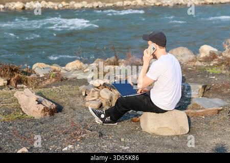 Ein junger Mann sitzt auf einem Felsen an einem Fluss und balanciert einen Laptop auf den Knien, während er mit seinem Telefon spricht, umgeben von Kieselsteinen und Herbstgras. Stockfoto