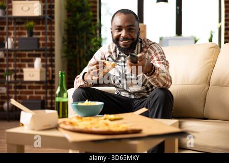 Happy Black man hält Pizzascheiben und Fernbedienung, wodurch die Lautstärke des Fernsehers erhöht wird, während er gute Filme genießt. Entspannter Wochenendmoment in einem modernen Apartment mit gemauerter Mauer, voller Komfort und Freude. Stockfoto