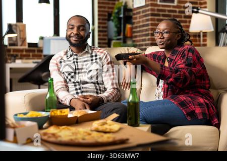 Schwarze Ehepartner schauen fern und essen Junk Food, fühlen sich entspannt und trinken zu Hause Flaschen Bier. Freund und Freundin saßen auf der Couch, während Frau die Kanäle wechselt und zufällige fernsehsendungen genossen. Stockfoto