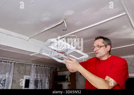 Installations-Elektriker hebt den neuen LED-Kronleuchter zur Decke, freiliegende Verkabelung sichtbar, Befestigung auf Befestigungspunkt ausgerichtet, Arbeiten werden ausgeführt. Stockfoto