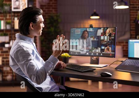 Fröhlicher Mann, der während eines Videoanrufs dem Computer zuwinkt, an Online-Meetings und Schulungen von zu Hause aus teilnimmt. Zuversichtlicher Remote-Mitarbeiter, der an virtuellen Gesprächen mit Kollegen und Kunden teilnimmt. Stockfoto