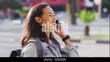 Sprechende asiatische Frau auf dem Smartphone am City plaza, mit Jeansjacke, Smartwatch, wachsamen Overlays Stockfoto