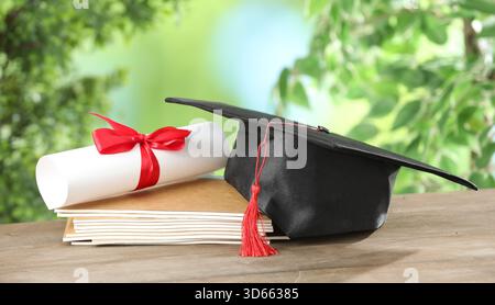 Graduierungskappe, Diplom und Notizbücher auf Holztisch vor verschwommenem Hintergrund, Großaufnahme Stockfoto