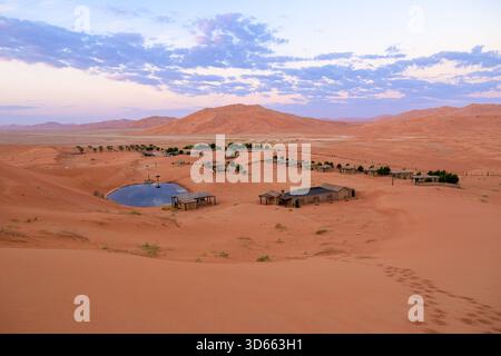 Abgelegenes Wüstencamp mit Zelten und Gebäuden rund um einen erfrischenden Oasenteich, eingebettet zwischen riesigen Sanddünen im leeren Viertel Oman Stockfoto