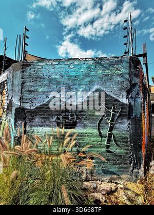 Wandgemälde auf einer alten Backsteinmauer, die eine bergige Landschaft mit Schiff und Figur darstellt, umgeben von wilden Gräsern unter einem lebendigen Himmel Stockfoto