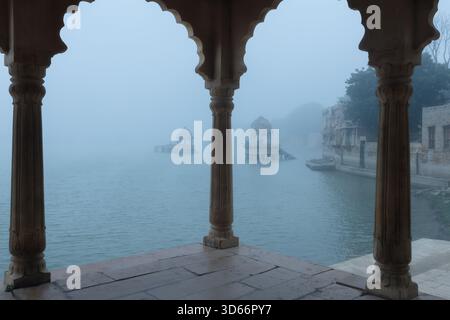 Nebliger Blick auf den Gadisar Lake in Jaisalmer, Rajasthan, mit alten Chhatris, die durch dichten Nebel unter einem Steinpavillon erscheinen. Stockfoto