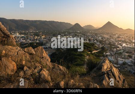 Aus der Vogelperspektive von Pushkar bei Sonnenuntergang zeigt die Stadt eingebettet zwischen felsigen Hügeln mit warmem Abendlicht, das die Landschaft von Rajasthan, Indien, beleuchtet Stockfoto