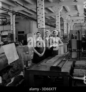 Slawjansk, Ukraine, UdSSR - Januar 1983: Porträt der Archivgruppe von drei jungen Arbeiterinnen, die an der Produktionslinie der Slawjansk-Bleistiftfabrik stehen. Das Schwarzweiß-Bild zeigt eine Brigade der Verpackungsabteilung mit Schürzen. Ein einzigartiges Detail sind die Betonsäulen, die an Birkenbäume erinnern und dem industriellen Innenraum einen Hauch von Natur verleihen. Dieses Foto veranschaulicht die Arbeitsbedingungen und die sowjetische Fabrikästhetik in der Region Donezk in den frühen 1980er Jahren Stockfoto