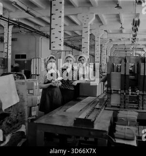 Slawjansk, Ukraine, UdSSR - Januar 1983: Porträt von drei Arbeiterinnen in der Slawjansk-Bleistiftfabrik. Die Brigade posiert in der Nähe eines Förderbandes, eine Frau hält einen Päckchen Bleistifte. Die Werkstatt verfügt über Säulen, die wie Birkenbäume bemalt sind, ein einzigartiger Versuch der industriellen Einrichtung. Dieses Schwarzweiß-Bild fängt die authentische Atmosphäre der sowjetischen Arbeit und die tägliche Routine einer Leichtindustrie im Winter in der Region Donezk ein. Stockfoto