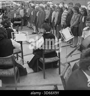 Slawjansk, Ukraine, UdSSR - 8. September 1985: Archivfoto des Stadttags mit einer kulturellen Massenvorstellung. Ein Kammerorchester begleitet einen großen Kinderchor von Mädchen mit weißen Bögen, die in Reihen stehen. Das Schwarz-weiß-Bild fängt die festliche Atmosphäre und die sowjetischen zeremoniellen Traditionen in der Region Donezk ein. Hinter den Musikern sind ein Wohngebäude und ein Bus zu sehen, die die authentische städtische Landschaft der friedlichen Ära widerspiegeln. Stockfoto