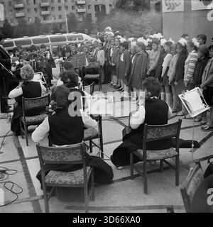 Slawjansk, Ukraine, UdSSR - 8. September 1985: Archivfoto des Stadttags mit einer kulturellen Massenvorstellung. Ein Kammerorchester begleitet einen großen Kinderchor von Mädchen mit weißen Bögen, die in Reihen stehen. Das Schwarz-weiß-Bild fängt die festliche Atmosphäre und die sowjetischen zeremoniellen Traditionen in der Region Donezk ein. Hinter den Musikern sind ein Wohngebäude und ein Bus zu sehen, die die authentische städtische Landschaft der friedlichen Ära widerspiegeln. Stockfoto