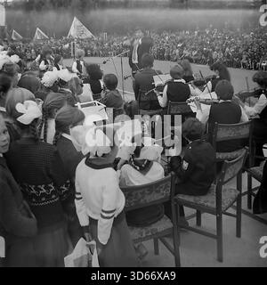 Slawjansk, Ukraine, UdSSR - 8. September 1985: Atmosphärisches Archivfoto, aufgenommen von der Bühne während des Stadtfestes. Ein männlicher Dirigent führt ein Streichorchester für Kinder, das für eine riesige Menge von Bürgern spielt, die sich auf dem Platz versammelt haben. Im Vordergrund sieht eine Gruppe von Mädchen mit großen weißen Bögen den Auftritt. Das Schwarzweiß-Bild fängt das Ausmaß des Feiertags und das kulturelle Leben der Donezker Region in der friedlichen Sowjetzeit ein. Stockfoto