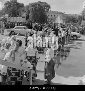 Slawjansk, Ukraine, UdSSR - 8. September 1985: Historisches Archivfoto des Stadtfestes auf dem zentralen Platz. Ein Volkstanz-Ensemble in traditionellen ukrainischen Kostümen begrüßt das Brautpaar, das in einem dekorierten Wolga-Auto ankommt. Das Schwarzweiß-Bild zeigt eine feierliche Zeremonie mit einer Braut und einem Bräutigam während einer öffentlichen Großveranstaltung. Im Hintergrund sind eine große Menschenmenge und ein Brunnen zu sehen, die die kulturellen Traditionen der Donezker Region in der Sowjetzeit widerspiegeln. Stockfoto
